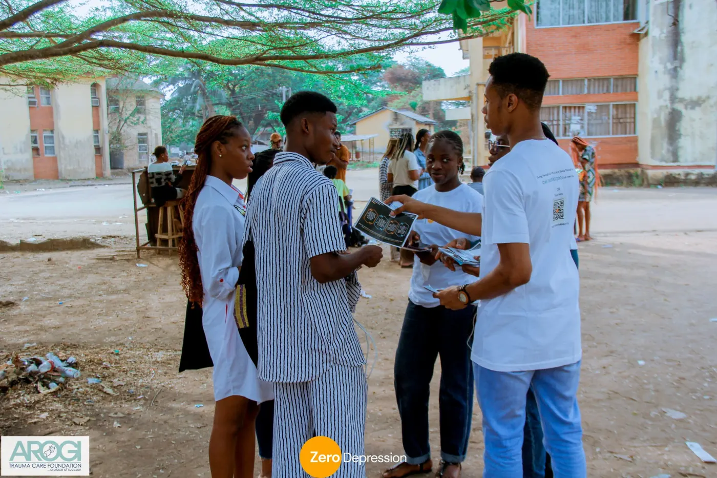 Two volunteers in white t-shirts handing out flyers to students under a large tree on campus